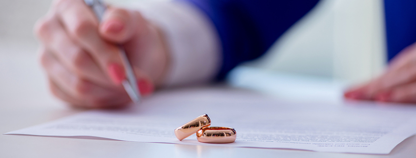 Person with painted nails signing document with wedding bands sitting nearby. Prenuptial Agreement