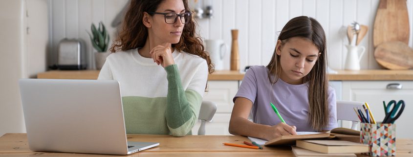Mother working remotely at home while her daughter does homework beside her at the kitchen table, showing a balance of parenting and remote work.