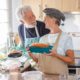 Senior couple in a domestic partnership smiling in their kitchen as the woman holds a homemade plum cake while her partner washes dishes in the background.