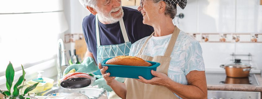 Senior couple in a domestic partnership smiling in their kitchen as the woman holds a homemade plum cake while her partner washes dishes in the background.