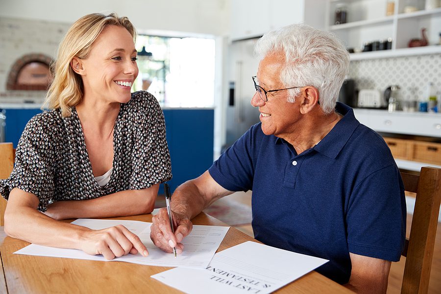 Woman assisting an elderly man at home as he fills out his last will and testament, symbolizing planning and family support.