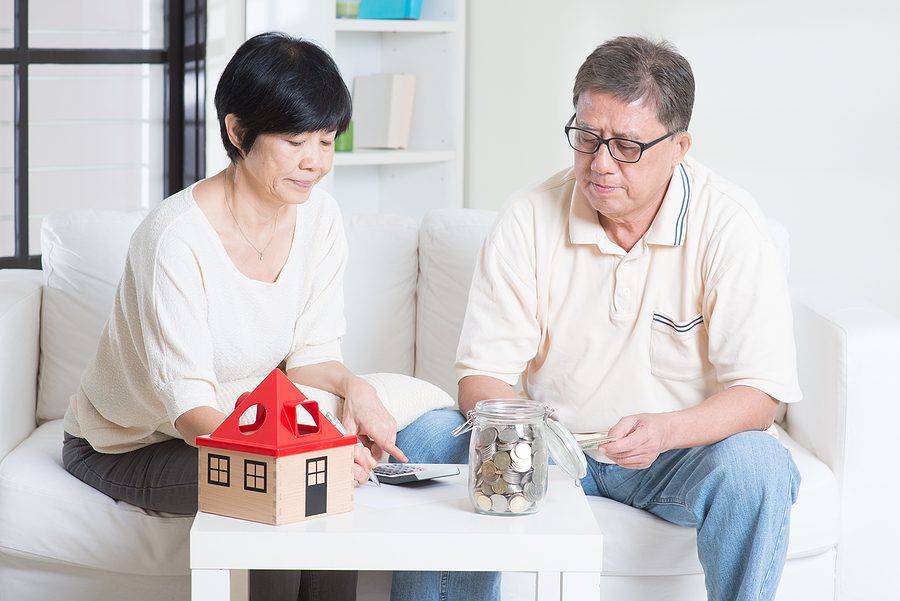 Asian senior couple planning retirement finances at home, counting savings and reviewing expenses beside a small house model.