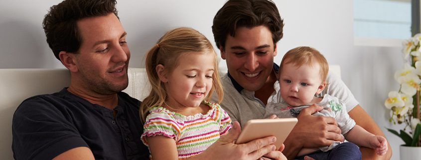 Two dads relaxing in bed with tablet and kids, an example of nontraditional family estate planning